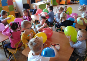Dzieci z grupy Wiewiórek siedzą przy stolikach w sali. Za pomocą mazaków ozdabiają nadmuchane, kolorowe balony.